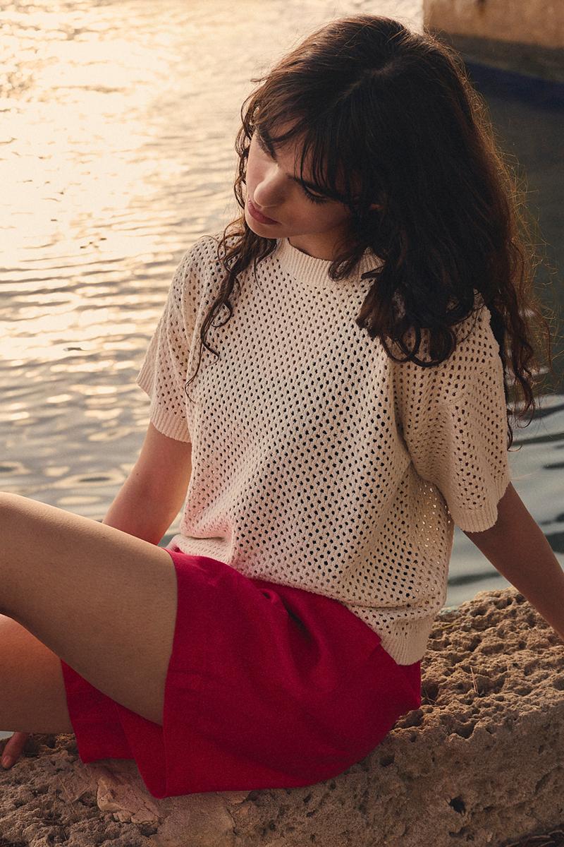 Eine Frau in einem cremefarbenen Häkel-Oberteil und pinkfarbenen Shorts von BITTE KAI RAND, die bei Sonnenuntergang auf einem Felsen am Wasser sitzt.