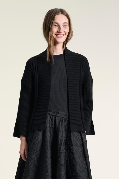 A smiling woman wearing a black BITTE KAI RAND cardigan with ribbed detailing, styled with a dark top and patterned skirt.